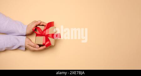 Die Hand einer Frau hält eine Geschenkbox, die in ein rotes Seidenband auf beigefarbenem Hintergrund gewickelt ist, von oben Stockfoto