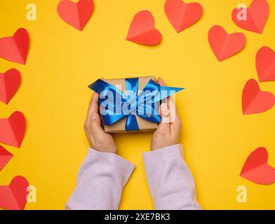 Die Hand einer Frau hält eine Geschenkbox in einem blauen Seidenband auf gelbem Hintergrund Stockfoto