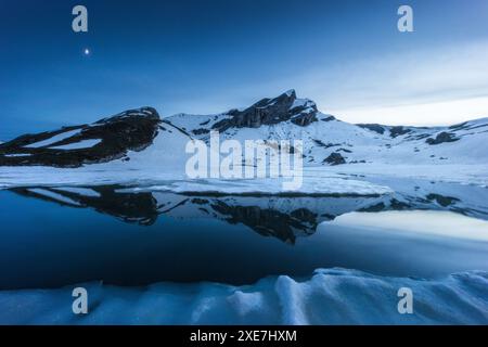 Ein malerischer Blick auf einen schneebedeckten Berg, der sich in einem teilweise gefrorenen See in Etang de Trente Pas, Wallis, Schweiz spiegelt Stockfoto