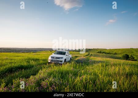 Weißer Volkswagen Amarok in einer hügeligen Landschaft. Vorderansicht des VW-Pickup-Trucks auf einer Wiese, der auf die Kamera zufährt. Stockfoto