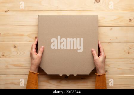 Pizza geschlossener Karton in menschlichen kaukasischen Händen auf natürlichem Holztisch flach liegendes Modell mit Rohling Stockfoto