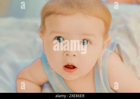 Der süße Junge mit blauen Augen und blonden Haaren liegt auf dem Bauch auf einer weichen Decke und sieht überrascht und neugierig aus, als er nach oben blickt Stockfoto