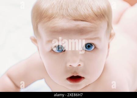 Der Junge mit blauen Augen und blonden Haaren liegt auf einer weißen Decke und blickt mit neugierigem und unschuldigem Ausdruck überraschend auf Stockfoto