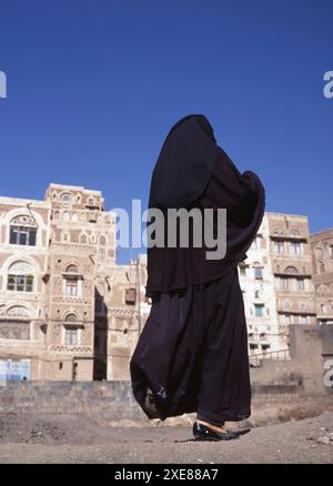 SANA'A, JEMEN - 23. APRIL 2011: Eine verschleierte muslimische Frau läuft auf einer Sana'a Straße im Jemen. Im Hintergrund typische jemenitische Häuser. Stockfoto