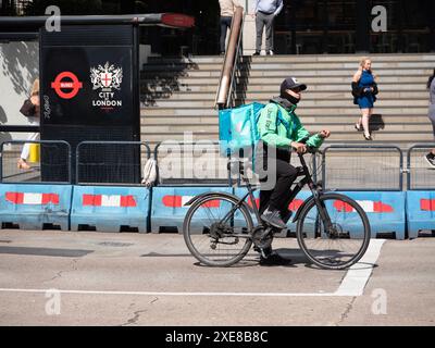 Ein Fahrer mit zwei Agenten, der eine Ober Eats Jacke mit Deliveroo Rucksack trägt, wartet an der Kreuzung in Bishopsgate, neben dem City of London Schild Stockfoto