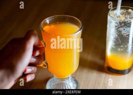 Eine Hand, die einen mit Orangensaft gefüllten Glasbecher hält, mit einem weiteren Glas Orangensaft mit Eis im Hintergrund auf einem Holztisch. Stockfoto