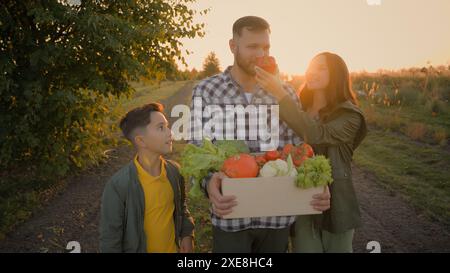 Glückliche kaukasische Familie Rückkehr nach der Landwirtschaft Arbeit ernten Mann männlicher Kerl Vater Tragen Kiste Gemüse kleines Kind Sohn Junge spazieren Frau Stockfoto