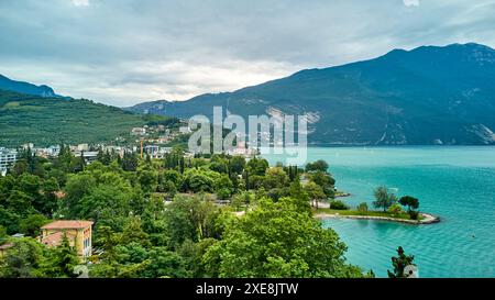 Riva del Garda, Gardasee, Italien - 25. Juni 2024: Zeigt den malerischen Gardasee, umgeben von üppigen grünen Wäldern und hohen Bergen, die eine beeindruckende Kulisse bieten. Charmante Gebäude und gepflegte Grünflächen laden zum Flanieren und Entspannen ein. *** Zeigt den malerischen Gardasee, umgeben von üppig grünen Wäldern und hohen Bergen, die eine beeindruckende Kulisse bieten. Am Ufer sind charmante Gebäude und gepflegte Grünanlagen zu sehen, die zu Spaziergängen und Erholung einladen. Stockfoto