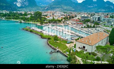 Riva del Garda, Gardasee, Italien - 25. Juni 2024: Kleiner Hafen von Riva del Garda mit Segelbooten und Segelschiffen und Blick vom Gardasee zu den Gebäuden der Stadt *** kleiner Hafen von Riva del Garda mit Segelbooten, bzw. Segelschiffen und Blick vom Gardasee auf die Gebäude der Stadt Stockfoto