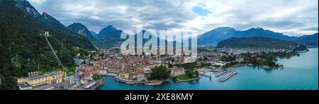 Riva del Garda, Gardasee, Italien - 25. Juni 2024: Das Panorama zeigt die Stadt Riva del Garda am nördlichen Ende des Gardasees, umgeben von majestätischen Bergen. Die Altstadt mit ihren charakteristischen, farbenfrohen Gebäuden erstreckt sich entlang der Küste, während der Hafen voller Boote und Yachten ist. Das grüne Laub der Bäume und das türkisfarbene Wasser des Sees schaffen eine malerische Szene, ergänzt durch die dramatischen Bergketten im Hintergrund. Der Himmel ist teilweise bewölkt und trägt zur atmosphärischen Tiefe des Bildes bei. *** Das Panorama zeigt die Stadt Riva del Garda am nördlic Stockfoto