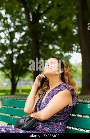 Lächelnde hispanische Frau, die auf einer Bank in einem Park in Paris sitzt und am Telefon spricht. Kopierbereich Stockfoto