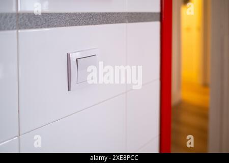 Detail eines weißen Lichtschalters an der gefliesten Wand einer Küche. Es gibt einen Raum, der im Hintergrund mit warmem Licht beleuchtet wird Stockfoto