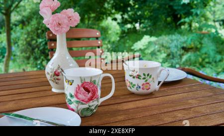 Gedeckter Tisch mit zwei hübschen Tassen, Tellern, einer Vase mit einer Rose auf einem hölzernen Gartentisch. Stockfoto