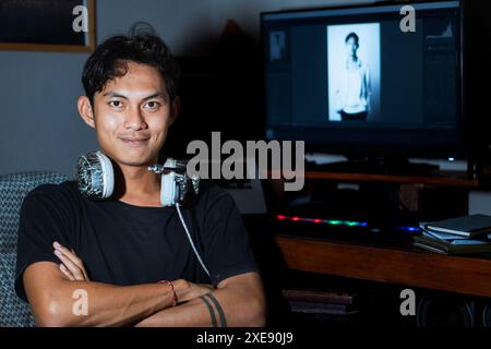 Ein junger Mann mit Kopfhörern um den Hals sitzt selbstbewusst vor einem Computer und zeigt ein Schwarzweißporträt auf dem Bildschirm. Stockfoto