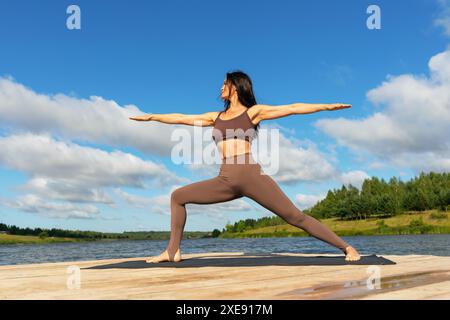 Frau mittleren Alters, die Virabhadrasana-Übungen durchführt, Kriegerstellung Nummer zwei, Yoga in Sportbekleidung am Ufer eines Sees auf einem warmen sonnigen Mo praktiziert Stockfoto