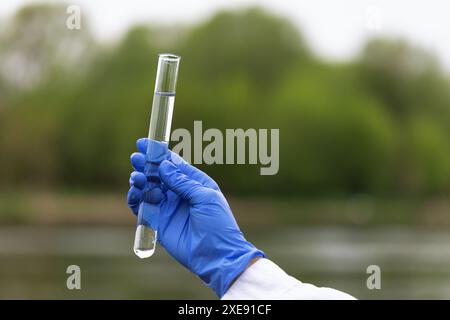 Nahaufnahme einer Hand in einem blauen Handschuh, die ein Glasröhrchen mit Flusswasserproben zur Analyse hält, ein Konzept zum Thema Wasserqualitätsprüfung Stockfoto