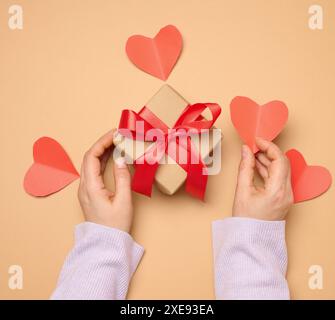 Die Hand einer Frau hält eine Geschenkbox in einem roten Seidenband auf beigefarbenem Hintergrund Stockfoto