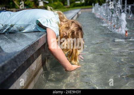 Ein junges Mädchen mit langen, blonden Haaren beugt sich über den Rand eines Brunnens und streckt ihre Hand ins Wasser. Das Wasser ist funkelnd und klar, und das Wasser ist klar Stockfoto