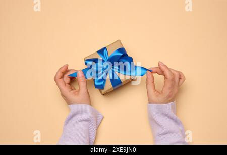 Die Hand einer Frau hält eine Geschenkbox in einem blauen Seidenband auf beigefarbenem Hintergrund Stockfoto