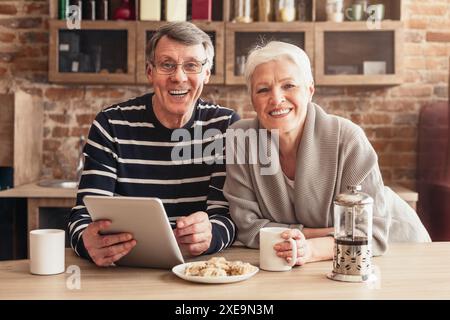 Glückliches Seniorenpaar, das Kaffee und Tablet in der modernen Küche genießt Stockfoto