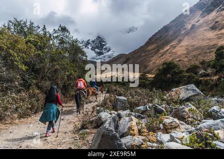 Lateinische Frau, die an einem sonnigen Tag mit Wolken und blauem Himmel in den Anden Perus auf einem Weg geht, umgeben von schneebedeckten Bergen und grüner Vegetation Stockfoto