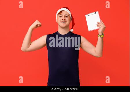 Porträt des persönlichen Trainers im Weihnachtsmann-Hut mit Klemmbrett mit Trainingsplan auf rotem Hintergrund Stockfoto