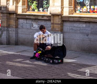 Straßenmusiker spielt Gitarre an einem sonnigen Tag in einer urbanen Umgebung, mit farbenfroher Geschenkbox und kopfsteingepflasterter Straße Falmouth Cornwall Stockfoto