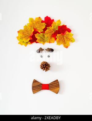 Lustiges und niedliches Jungen Gesicht gemacht mit bunten Herbstblättern, Nüssen, Tannenzapfen und hölzernen Schleife auf weißem Hintergrund. Minimales Herbst- oder Herbstkonzept. Stockfoto