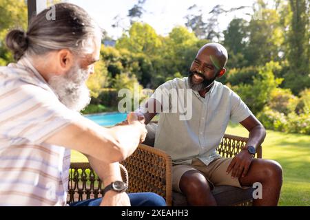 Ältere Freunde schütteln die Hände und lächeln, während sie draußen im Garten sitzen Stockfoto