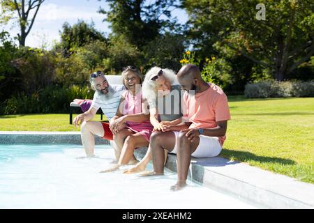 Eine Gruppe älterer Freunde sitzt am Pool, genießt den sonnigen Tag und lacht Stockfoto