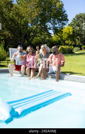 Mit Getränken toasten, verschiedene Seniorenfreunde sitzen am Pool und genießen den Sommer Stockfoto