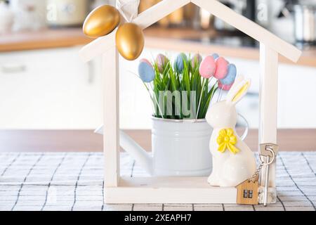 Schlüssel und winziges Haus mit gemütlichem Haus mit Osterdekor mit Kaninchen und Eiern auf dem Tisch der Küche. Gebäude, Design, Projekt, Umzug nach Stockfoto