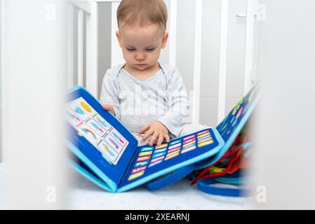 Baby spielt mit montessori ruhigem Buch, das in einem Kinderbett sitzt. Das Konzept von Smart Books und modernem Lernspielzeug Stockfoto