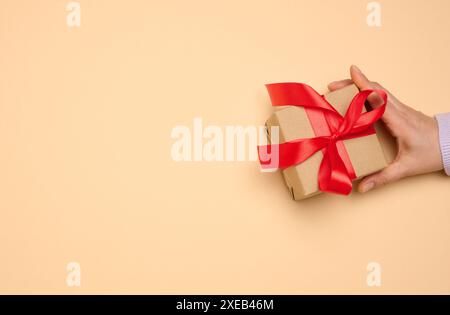 Die Hand einer Frau hält eine Geschenkbox in einem roten Seidenband auf beigefarbenem Hintergrund Stockfoto