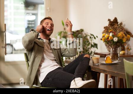 Lifestyle-Porträt eines hübschen jungen Mannes, der im Café sitzt, mit einem Freund auf dem Handy mit schnurlosen Ohrhörern spricht, lacht Stockfoto