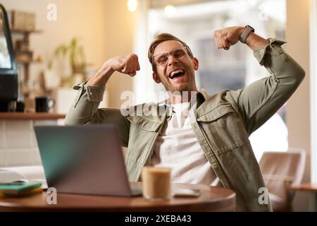 Porträt des gutaussehenden, zufriedenen jungen Mannes zeigt Bizeps, beugt Muskeln, sitzt vor dem Laptop im Café, freut sich, privater Trainer Stockfoto