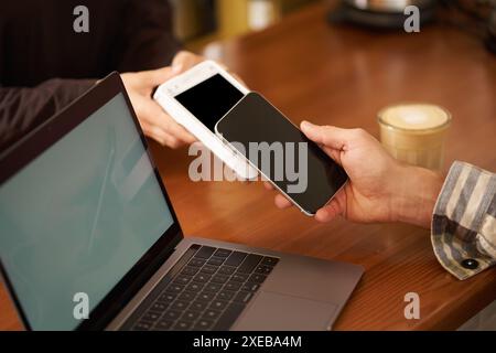 Zugeschnittene Aufnahme, in der das Telefon per Hand an das POS-Zahlungsterminal gedrückt wird, um kontaktlos für eine Bestellung zu bezahlen, wobei das Smartphone zum Einkaufen verwendet wird Stockfoto