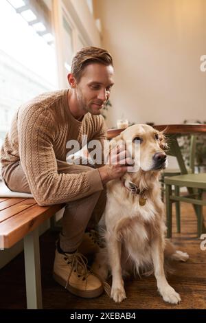 Nahporträt eines gutaussehenden Mannes mit seinem Hund, der im Café sitzt. Ein Typ trinkt seinen Kaffee und berührt Golden Retriever Stockfoto