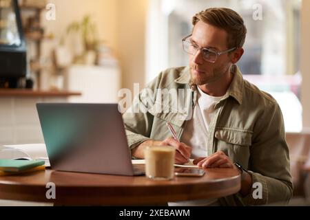 Porträt eines gutaussehenden jungen Mannes in Brille, Mann, der lernt, Notizen macht, während des Online-Unterrichts auf den Laptop-Bildschirm schaut, nimmt an einer Teil Stockfoto