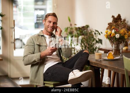 Lifestyle-Porträt eines hübschen, stilvollen jungen Mannes, sitzt im Café, hält das Handy, zieht schnurlose Ohrhörer an, schaut in die Kamera Stockfoto