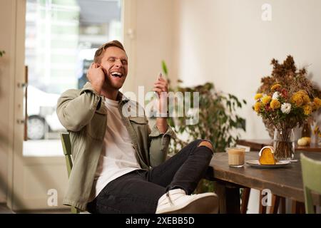 Lifestyle-Porträt eines hübschen jungen Mannes, der im Café sitzt, mit einem Freund auf dem Handy mit schnurlosen Ohrhörern spricht, lacht Stockfoto