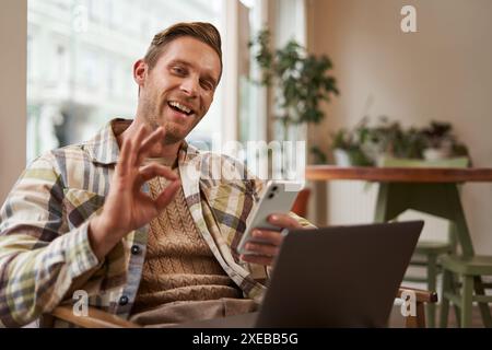 Porträt eines gutaussehenden Mannes im Café, sitzt mit Laptop, arbeitet ferngesteuert, freiberuflich mit Telefon in den Händen, zeigt okay, okay Geste Stockfoto
