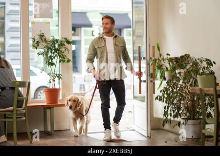 Porträt eines gutaussehenden, lächelnden jungen Mannes, der mit seinem süßen Hund in das Café geht, goldener Retriever an der Leine. Guy tritt in pe ein Stockfoto