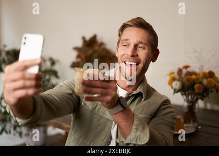 Fröhlicher, gutaussehender junger Mann, Selfie in der Handy-App, Video-Chats mit jemandem und ein Glas Kaffee heben, jubelt Gestu an Stockfoto