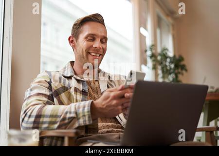 Nahaufnahme Porträt eines hübschen lächelnden Mannes mit Laptop, Café-Besucher, der auf Stuhl sitzt, Smartphone benutzt, glücklich aussieht, Nachrichten sendet Stockfoto