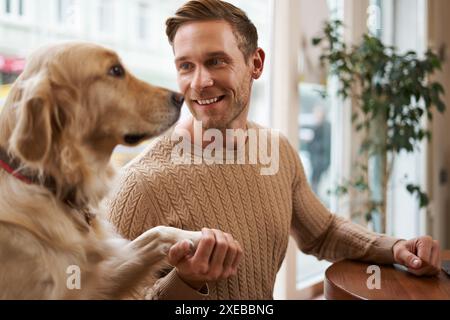 Nahaufnahme Porträt eines süßen, klugen Hundes gibt seinem Besitzer die Pfote. Schöner Mann mit seinem goldenen Retriever, der in einem haustierfreundlichen Café sitzt Stockfoto