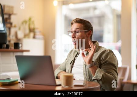 Porträt eines jungen Freelancers, eines digitalen Nomaden, der ein Online-Meeting hat, Video-Chats, zeigt OK-Zeichen am Laptop, bestätigt etwas, Stockfoto