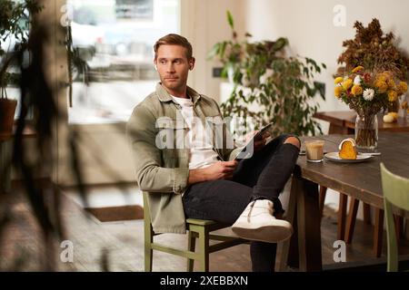 Lifestyle-Aufnahme eines hübschen jungen Mannes, der in einem Café vor dem Tisch sitzt, Kaffee trinkt, kabellose Kopfhörer trägt, Liste Stockfoto