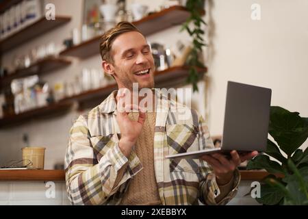 Porträt eines jungen lächelnden Mannes, ein Kollege nimmt an einem Online-Meeting vom Café Teil, Video-Chats auf Laptop, zeigt alles okay, bitte handsignieren Stockfoto