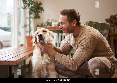 Porträt eines glücklichen lächelnden, gutaussehenden Mannes, streichelt seinen goldenen Retriever und lacht. Ein junger Kerl mit seinem Hund in einem Café Stockfoto